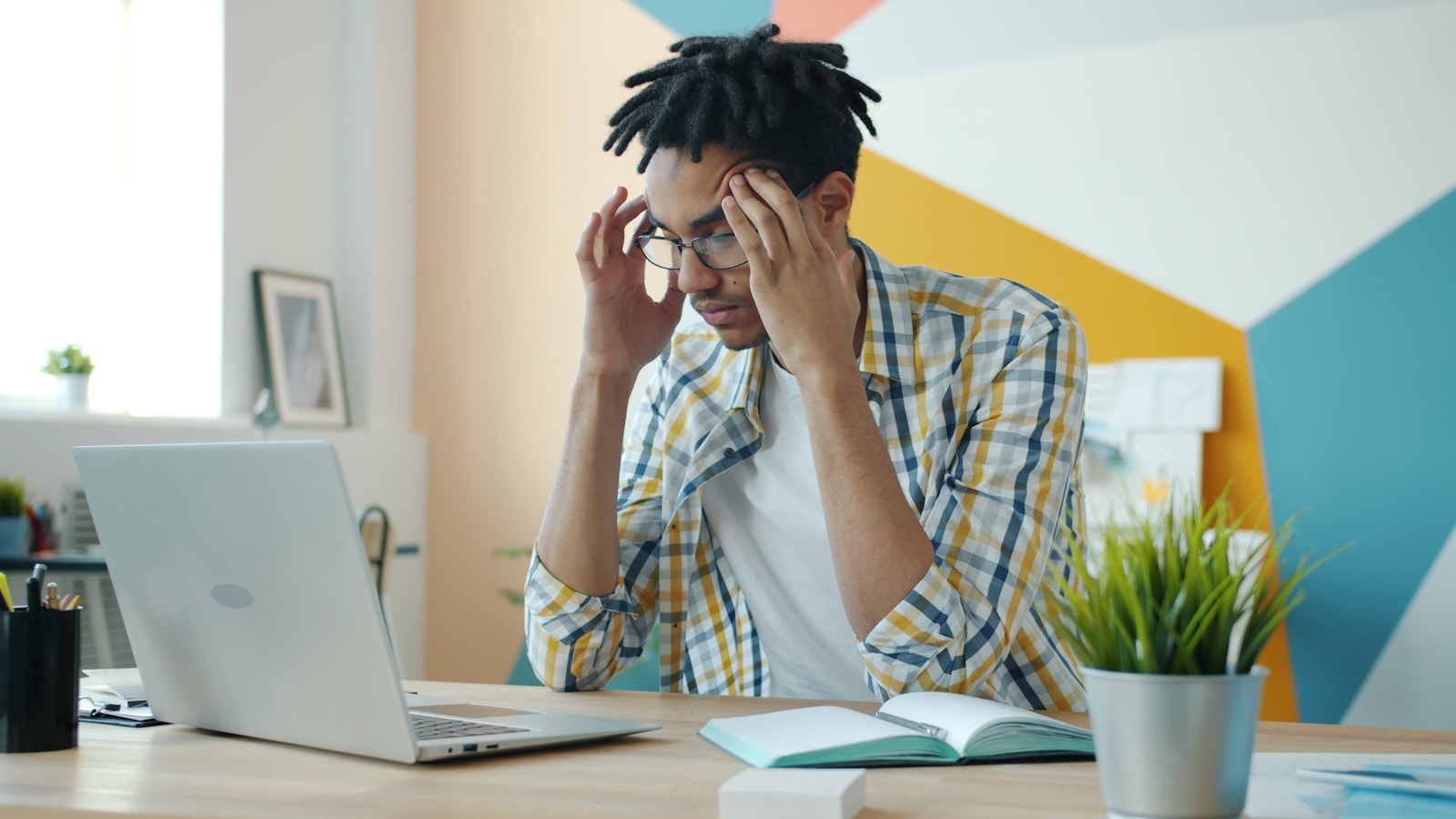 Man with dreadlocks holding head at desk with laptop